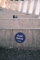 A circular blue sticker with the words 'Good Karma' and 'Rework' is affixed to a concrete surface. The background features a blurred, repetitive pattern suggesting steps or seating rows, likely in an outdoor setting. The ground in front of the concrete appears rough and slightly littered.