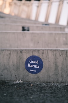 A circular blue sticker with the words 'Good Karma' and 'Rework' is affixed to a concrete surface. The background features a blurred, repetitive pattern suggesting steps or seating rows, likely in an outdoor setting. The ground in front of the concrete appears rough and slightly littered.