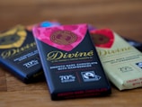 A close-up view of three chocolate bars laid on a wooden surface. The central bar has a vibrant pink and black wrapper with gold lettering that reads 'Divine' and is labeled as smooth dark chocolate with raspberries containing 70% cocoa. The nearby bars have varying wrapping designs, including a light-colored one labeled as creamy white chocolate with strawberries with 25% cocoa.