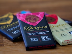 A close-up view of three chocolate bars laid on a wooden surface. The central bar has a vibrant pink and black wrapper with gold lettering that reads 'Divine' and is labeled as smooth dark chocolate with raspberries containing 70% cocoa. The nearby bars have varying wrapping designs, including a light-colored one labeled as creamy white chocolate with strawberries with 25% cocoa.