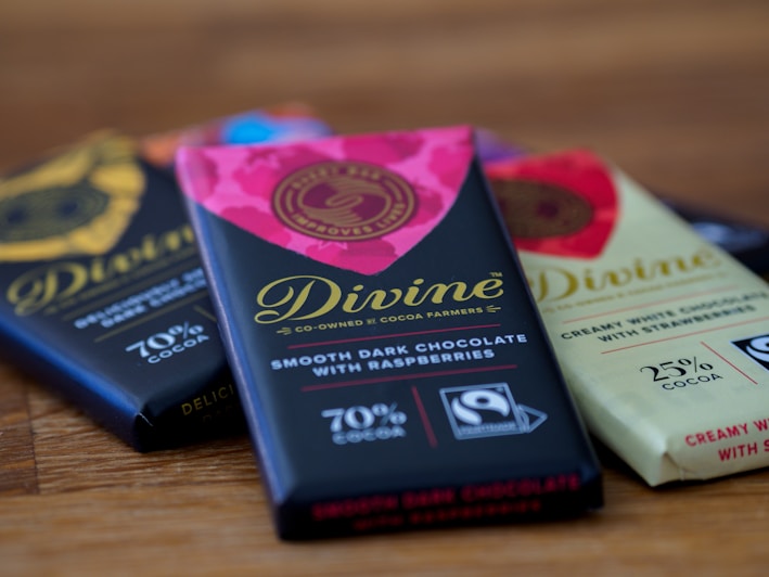 A close-up view of three chocolate bars laid on a wooden surface. The central bar has a vibrant pink and black wrapper with gold lettering that reads 'Divine' and is labeled as smooth dark chocolate with raspberries containing 70% cocoa. The nearby bars have varying wrapping designs, including a light-colored one labeled as creamy white chocolate with strawberries with 25% cocoa.