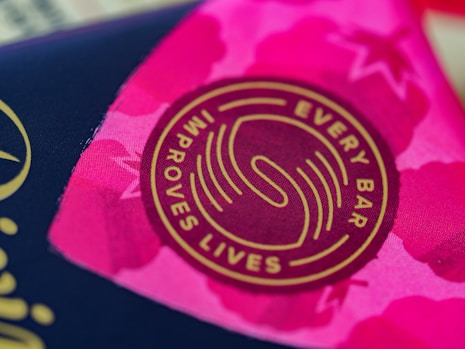 A close-up view of a circular logo with the words 'Every Bar Improves Lives' written around a stylized design. The background displays vibrant shades of pink with abstract patterns, possibly floral or star-like shapes.