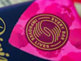 A close-up view of a circular logo with the words 'Every Bar Improves Lives' written around a stylized design. The background displays vibrant shades of pink with abstract patterns, possibly floral or star-like shapes.