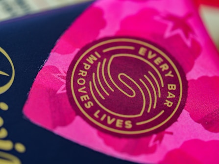 A close-up view of a circular logo with the words 'Every Bar Improves Lives' written around a stylized design. The background displays vibrant shades of pink with abstract patterns, possibly floral or star-like shapes.