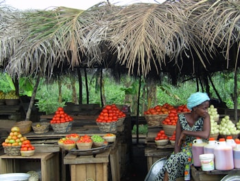 A rustic market stall displays an array of fresh produce, including baskets of tomatoes, citrus fruits, and green vegetables. A woman dressed in a colorful outfit and headscarf sits near the produce, looking to the side. The stall is covered with a thatched roof made of palm leaves, and the background features lush green foliage.