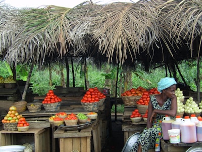 A rustic market stall displays an array of fresh produce, including baskets of tomatoes, citrus fruits, and green vegetables. A woman dressed in a colorful outfit and headscarf sits near the produce, looking to the side. The stall is covered with a thatched roof made of palm leaves, and the background features lush green foliage.