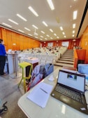 A lecture hall with rows of seats and people sitting, some wearing masks. A person is standing at a podium on the left, and there is a table with a laptop, papers, and other items at the foreground. The ceiling has bright lights, and the walls are wooden.