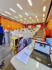 A lecture hall with rows of seats and people sitting, some wearing masks. A person is standing at a podium on the left, and there is a table with a laptop, papers, and other items at the foreground. The ceiling has bright lights, and the walls are wooden.