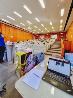 A lecture hall with rows of seats and people sitting, some wearing masks. A person is standing at a podium on the left, and there is a table with a laptop, papers, and other items at the foreground. The ceiling has bright lights, and the walls are wooden.