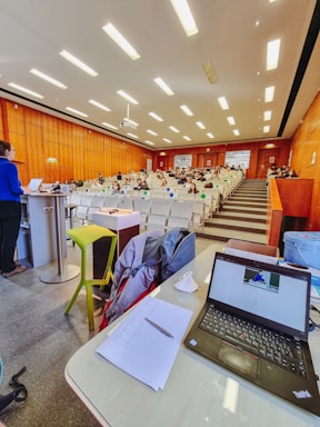 A lecture hall with rows of seats and people sitting, some wearing masks. A person is standing at a podium on the left, and there is a table with a laptop, papers, and other items at the foreground. The ceiling has bright lights, and the walls are wooden.