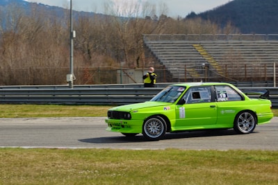 A bright green racing car is captured on a track with mountains and barren trees in the background. The car has various decals and sponsorships, and is driving past empty bleachers. A person in the background appears to be taking pictures or observing the race.