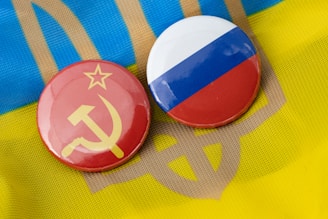 Two circular badges are displayed, one with the Soviet hammer and sickle on a red background, and the other featuring the Russian flag with horizontal stripes of white, blue, and red. They are placed on top of a textile with the colors and pattern resembling the Ukrainian flag.