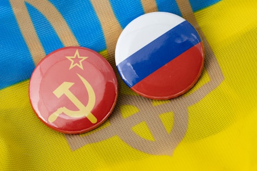 Two circular badges are displayed, one with the Soviet hammer and sickle on a red background, and the other featuring the Russian flag with horizontal stripes of white, blue, and red. They are placed on top of a textile with the colors and pattern resembling the Ukrainian flag.