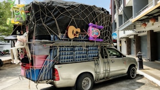 A pickup truck loaded with a variety of household items and furniture, secured with netting. The cargo includes a guitar, plastic bins, a mattress, and children's toys such as a tricycle and a baby walker. The truck is parked on a street adjacent to buildings and a tree, with some traditional lanterns hanging from a storefront.
