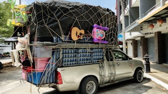 A pickup truck loaded with a variety of household items and furniture, secured with netting. The cargo includes a guitar, plastic bins, a mattress, and children's toys such as a tricycle and a baby walker. The truck is parked on a street adjacent to buildings and a tree, with some traditional lanterns hanging from a storefront.