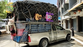 A pickup truck loaded with a variety of household items and furniture, secured with netting. The cargo includes a guitar, plastic bins, a mattress, and children's toys such as a tricycle and a baby walker. The truck is parked on a street adjacent to buildings and a tree, with some traditional lanterns hanging from a storefront.