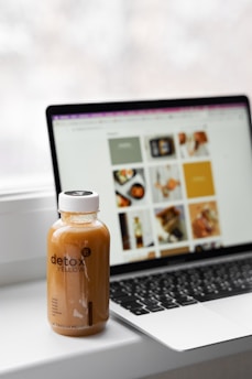 A bottle labeled 'detox yellow' is placed on a desk next to an open laptop. The laptop screen displays several images, most of which are blurred due to the focus on the bottle. The background appears to be bright, with natural light suggesting a window behind.