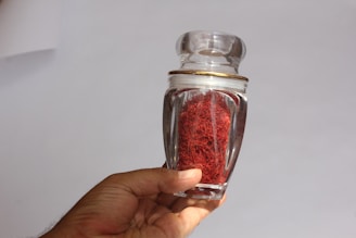 A hand holding a clear glass jar filled with red saffron threads against a plain background. The jar has a decorative gold rim on its lid.