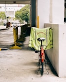 A red bicycle is parked against a white concrete wall, with a neon-green safety vest draped over the seat. The vest has the word 'SECURITY' printed in red capital letters. In the background, there's an outdoor parking area with yellow barriers and some trees visible further back.