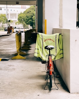 A red bicycle is parked against a white concrete wall, with a neon-green safety vest draped over the seat. The vest has the word 'SECURITY' printed in red capital letters. In the background, there's an outdoor parking area with yellow barriers and some trees visible further back.