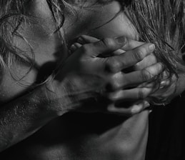 A black and white photograph focusing on hands and hair placed over a bare chest. The lighting emphasizes the texture of the skin and the strands of hair, creating dramatic shadows and highlights.