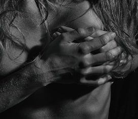 A black and white photograph focusing on hands and hair placed over a bare chest. The lighting emphasizes the texture of the skin and the strands of hair, creating dramatic shadows and highlights.