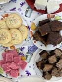Assorted baked goods are arranged on plates. There are colorful sprinkle cookies, dark chocolate brownies, pink coconut squares, and white coconut squares. The background is a tablecloth with floral and fruit patterns.