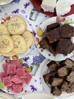 Assorted baked goods are arranged on plates. There are colorful sprinkle cookies, dark chocolate brownies, pink coconut squares, and white coconut squares. The background is a tablecloth with floral and fruit patterns.