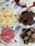 Assorted baked goods are arranged on plates. There are colorful sprinkle cookies, dark chocolate brownies, pink coconut squares, and white coconut squares. The background is a tablecloth with floral and fruit patterns.