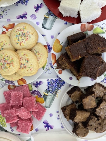 Assorted baked goods are arranged on plates. There are colorful sprinkle cookies, dark chocolate brownies, pink coconut squares, and white coconut squares. The background is a tablecloth with floral and fruit patterns.
