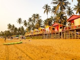A sandy beach lined with colorful elevated huts in a tropical setting, surrounded by tall palm trees. Some people can be seen walking or engaging in activities on the beach, with a clear sky above providing a warm and relaxing atmosphere.