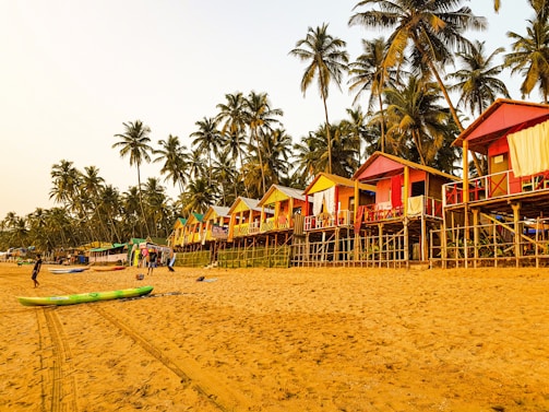 A sandy beach lined with colorful elevated huts in a tropical setting, surrounded by tall palm trees. Some people can be seen walking or engaging in activities on the beach, with a clear sky above providing a warm and relaxing atmosphere.
