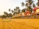 A sandy beach lined with colorful elevated huts in a tropical setting, surrounded by tall palm trees. Some people can be seen walking or engaging in activities on the beach, with a clear sky above providing a warm and relaxing atmosphere.