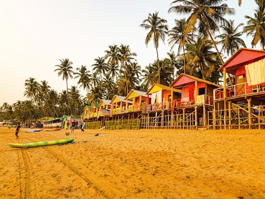 A sandy beach lined with colorful elevated huts in a tropical setting, surrounded by tall palm trees. Some people can be seen walking or engaging in activities on the beach, with a clear sky above providing a warm and relaxing atmosphere.