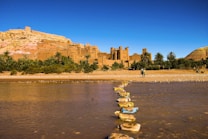 A historic desert city with earthen buildings set against a hilly backdrop under a clear blue sky. A river with stepping stones crosses the foreground, and lush greenery interspersed with palm trees surrounds the city. Two people are walking near the riverbank.
