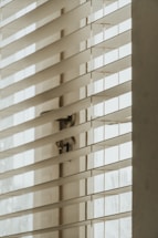 A close-up view of window blinds partially open, allowing soft natural light to filter through, creating shadows and a calm atmosphere.