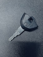 A single car key rests on a textured dark surface. The key has a black plastic head with a distinct shape and a metal blade exhibiting grooves specific to its locking mechanism.
