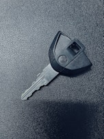 A single car key rests on a textured dark surface. The key has a black plastic head with a distinct shape and a metal blade exhibiting grooves specific to its locking mechanism.