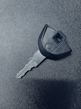 A single car key rests on a textured dark surface. The key has a black plastic head with a distinct shape and a metal blade exhibiting grooves specific to its locking mechanism.