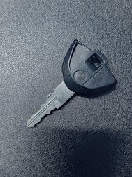 A single car key rests on a textured dark surface. The key has a black plastic head with a distinct shape and a metal blade exhibiting grooves specific to its locking mechanism.