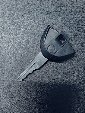 A single car key rests on a textured dark surface. The key has a black plastic head with a distinct shape and a metal blade exhibiting grooves specific to its locking mechanism.