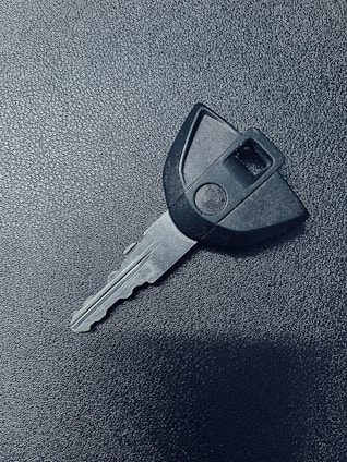 A single car key rests on a textured dark surface. The key has a black plastic head with a distinct shape and a metal blade exhibiting grooves specific to its locking mechanism.