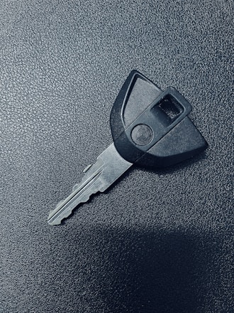 A single car key rests on a textured dark surface. The key has a black plastic head with a distinct shape and a metal blade exhibiting grooves specific to its locking mechanism.