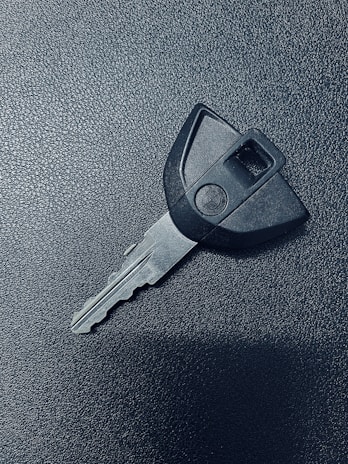 A single car key rests on a textured dark surface. The key has a black plastic head with a distinct shape and a metal blade exhibiting grooves specific to its locking mechanism.