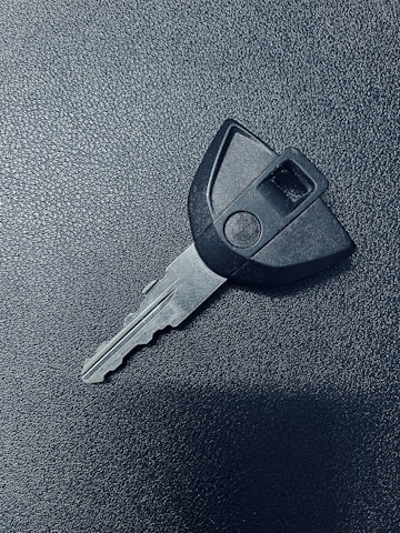 A single car key rests on a textured dark surface. The key has a black plastic head with a distinct shape and a metal blade exhibiting grooves specific to its locking mechanism.