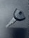 A single car key rests on a textured dark surface. The key has a black plastic head with a distinct shape and a metal blade exhibiting grooves specific to its locking mechanism.