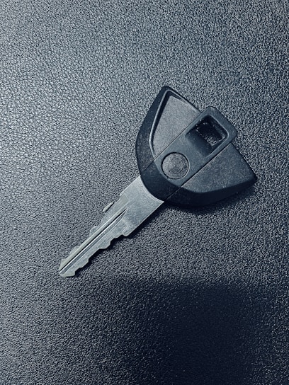 A single car key rests on a textured dark surface. The key has a black plastic head with a distinct shape and a metal blade exhibiting grooves specific to its locking mechanism.