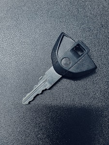 A single car key rests on a textured dark surface. The key has a black plastic head with a distinct shape and a metal blade exhibiting grooves specific to its locking mechanism.