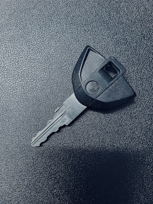 A single car key rests on a textured dark surface. The key has a black plastic head with a distinct shape and a metal blade exhibiting grooves specific to its locking mechanism.