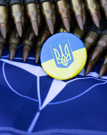 A button with the Ukrainian coat of arms and colors is placed on a blue background, possibly representing NATO. Surrounding the button are several bullets, arranged in a row, with their brass casings and pointed tips visible.